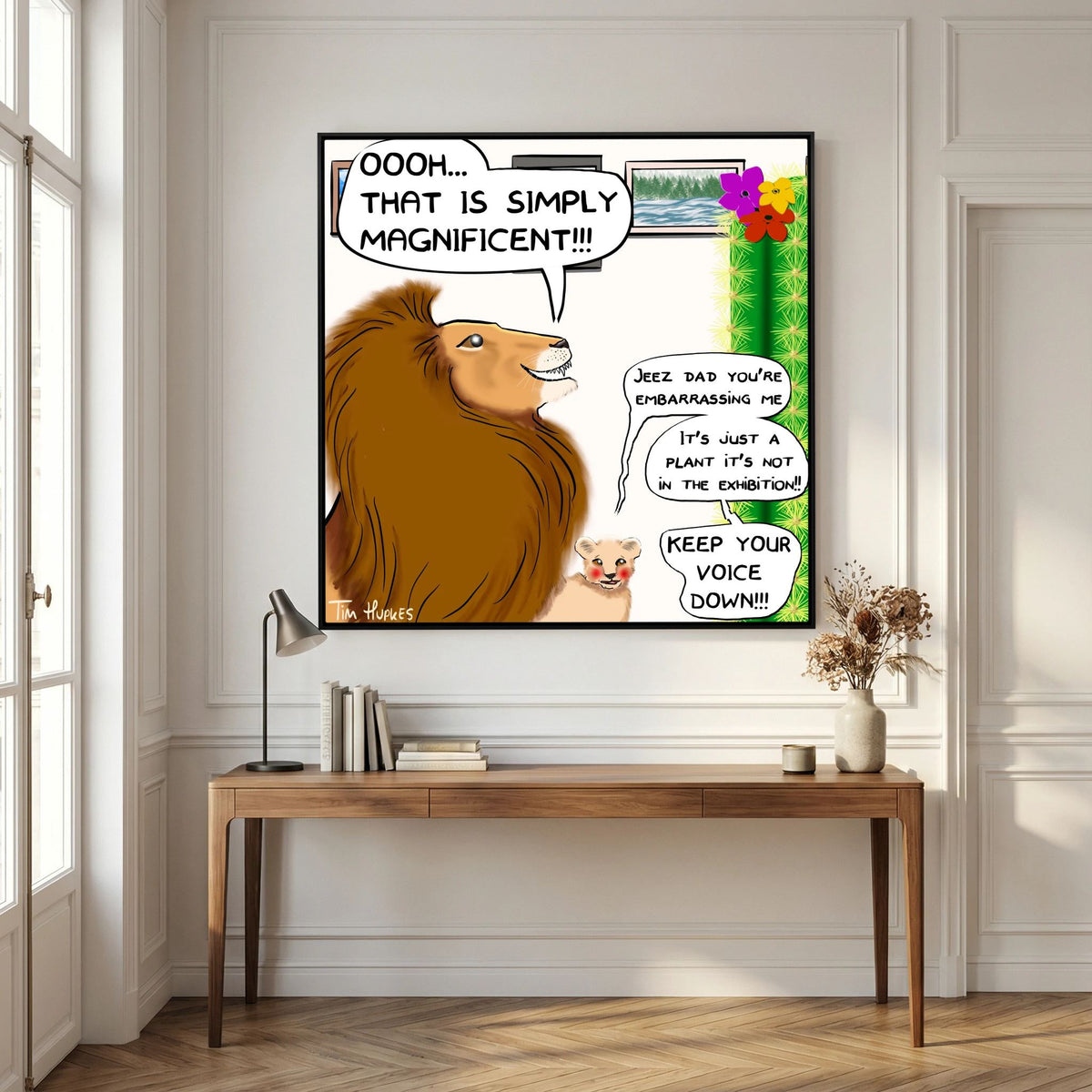 Black-framed Faux Pas by Tim Hupkes print of a lion and cub hanging above a wooden console table with a lamp and books