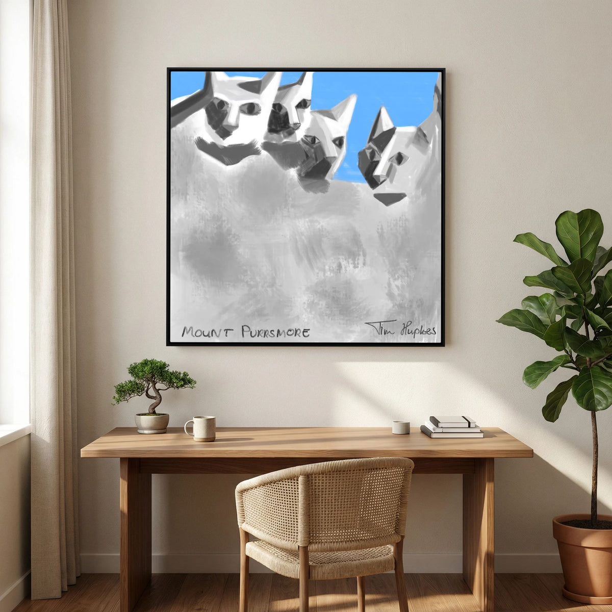 Black-framed Mount Purrsmore by Tim Hupkes canvas print above a wooden desk with a rattan chair, a bonsai and a large fiddle-leaf plant
