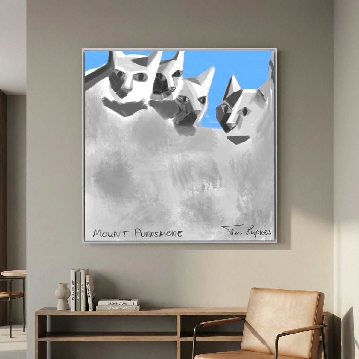 Mount Purrsmore by Tim Hupkes canvas print in a white frame above a wooden sideboard with books, beside a tan leather armchair