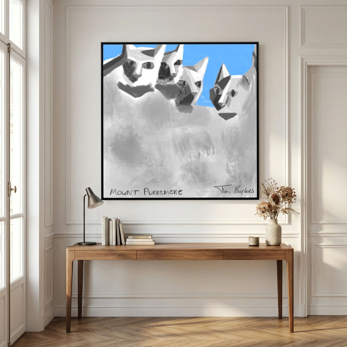 Black-framed Mount Purrsmore by Tim Hupkes canvas print on a panelled white wall above a wooden console table with a lamp and books